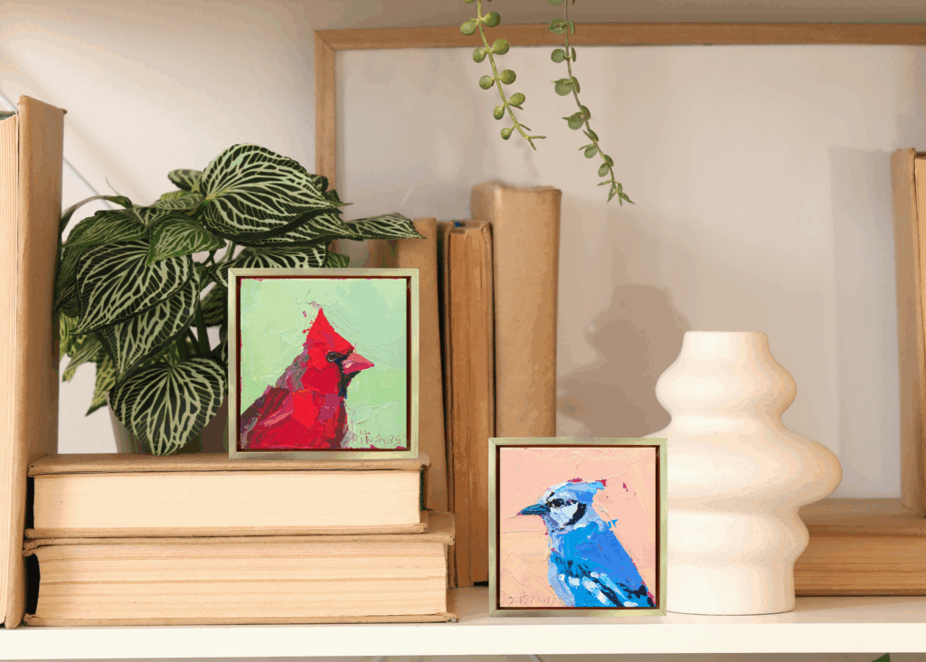 Two small framed acrylic bird paintings—a red cardinal and a blue jay—shown side by side on a shelf